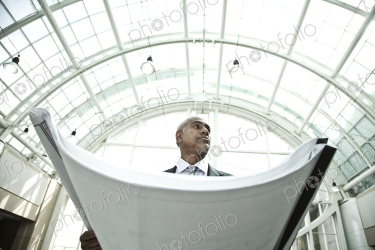 A businessman holding a large plan or architectural drawings standing under a glass…
