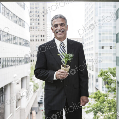 A businessman holding a small plant while stanidng next to a a large convention center… A businessman holding a small plant while stanidng next to a a large convention center…