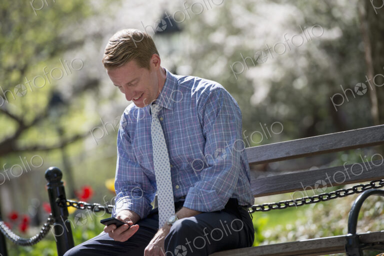 A businessman in a shirt with white tie, sitting on a park bench under the shade of a…