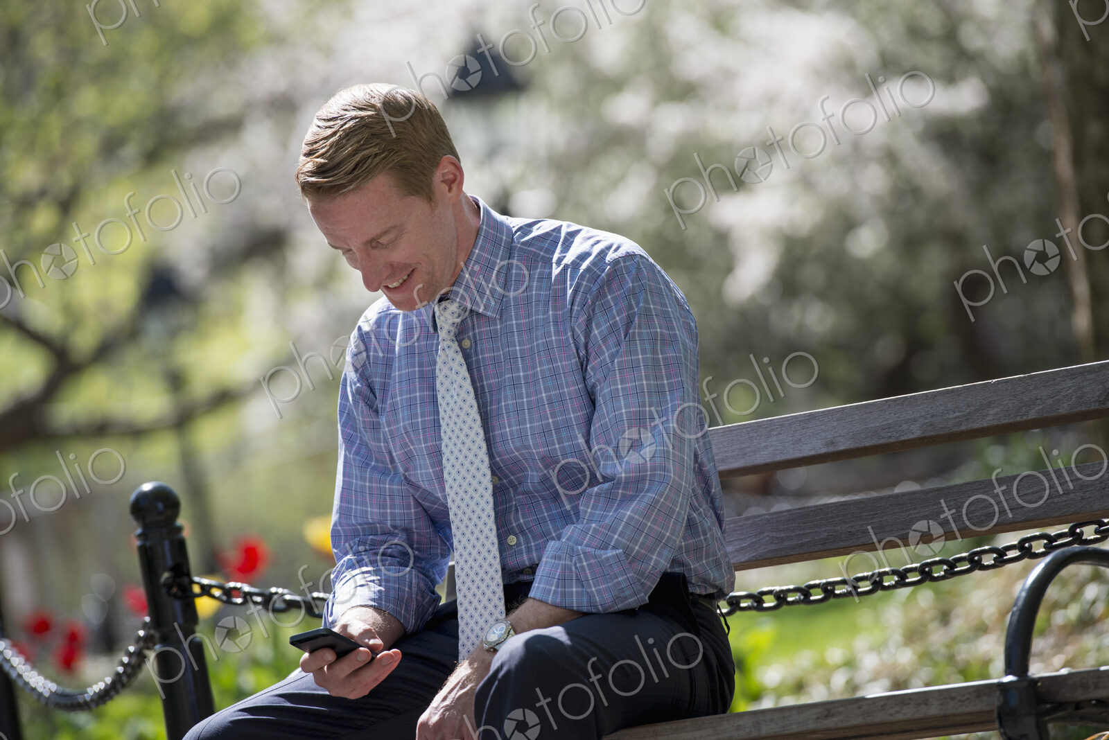 A businessman in a shirt with white tie, sitting on a park bench under the shade of a…