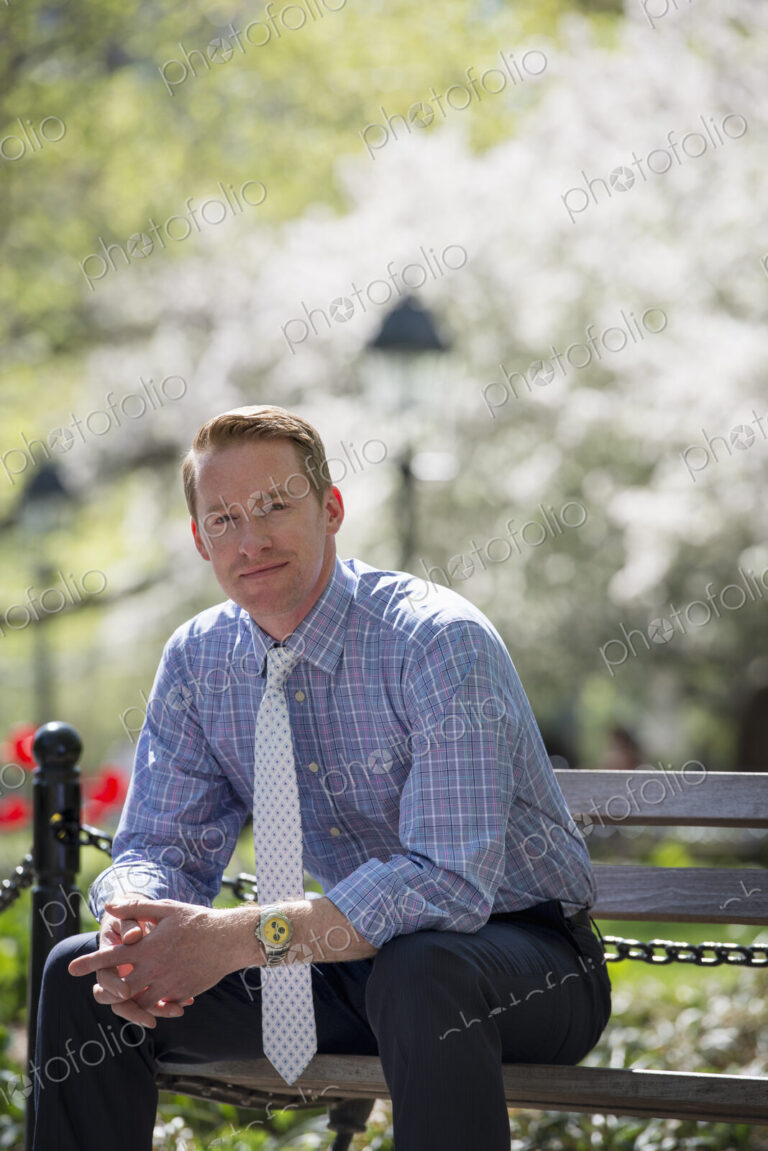 A businessman in a shirt with white tie, sitting on a park bench under the shade of a…