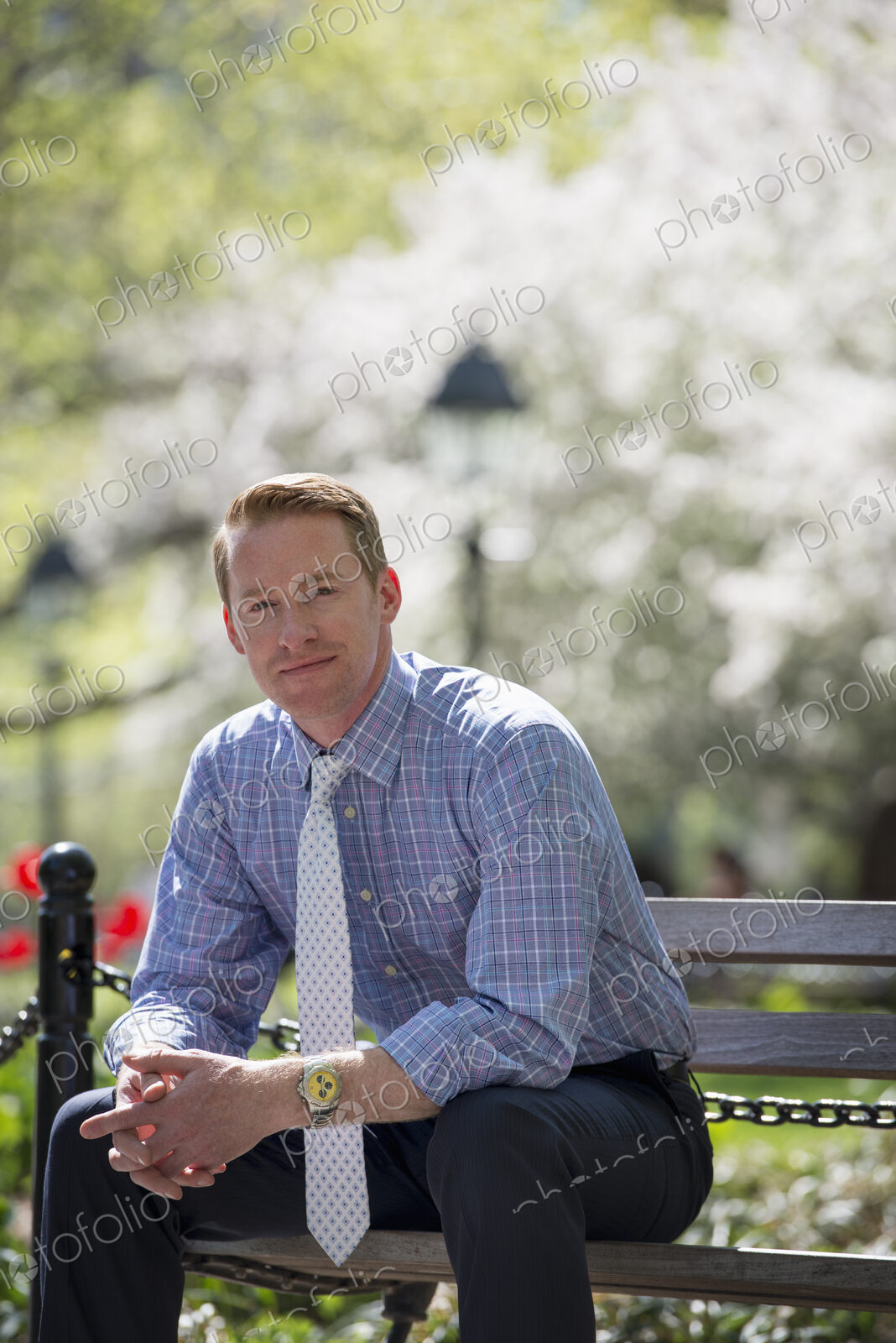 A businessman in a shirt with white tie, sitting on a park bench under the shade of a…