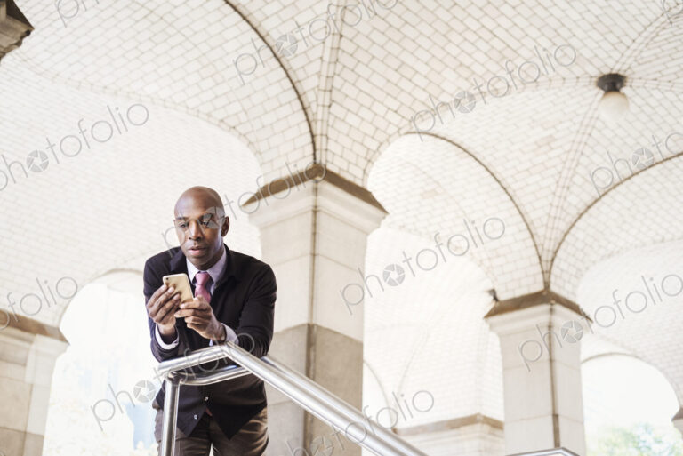 A businessman in a suit standing by a stairwell under an arched ceiling, checking his…