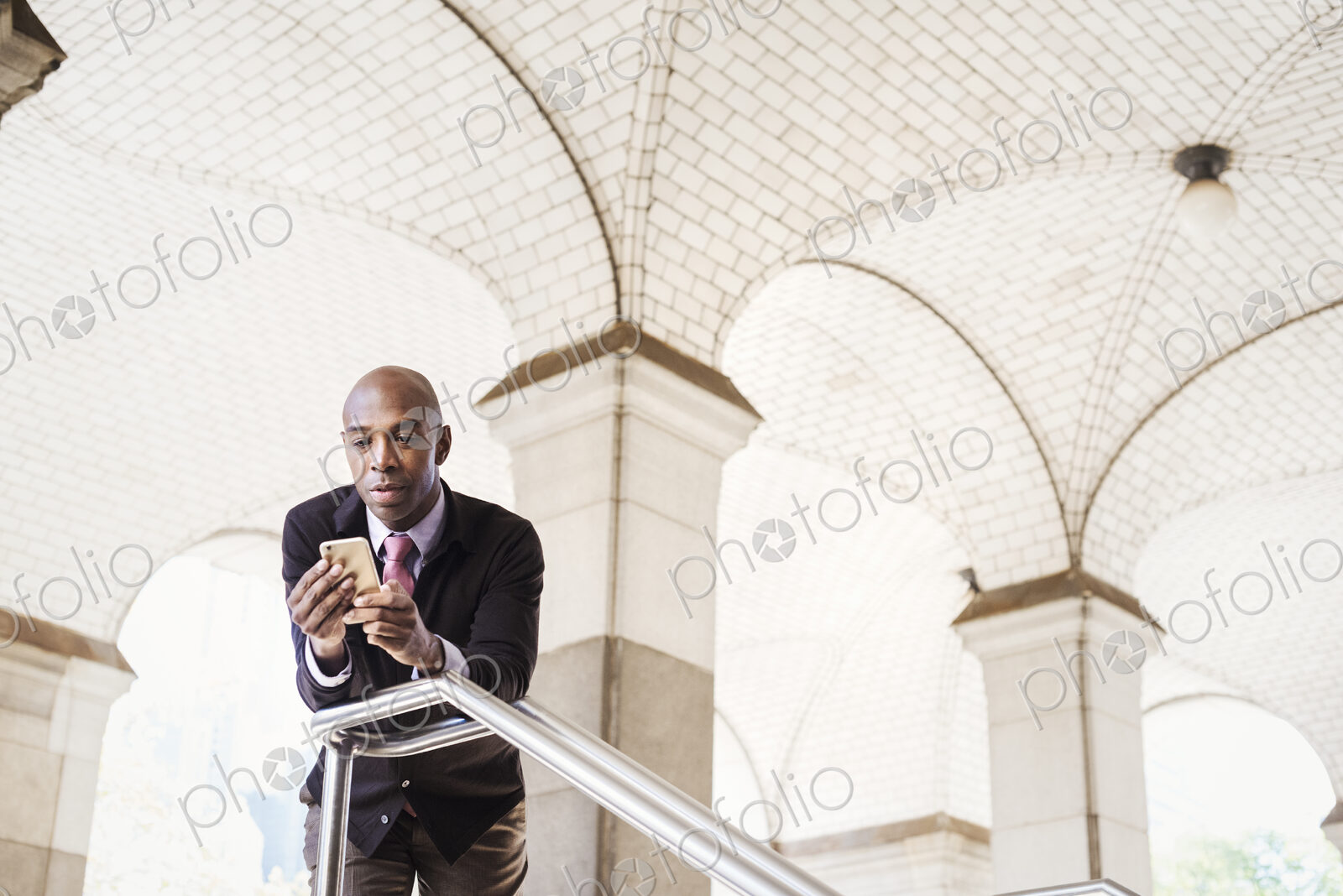 A businessman in a suit standing by a stairwell under an arched ceiling, checking his…
