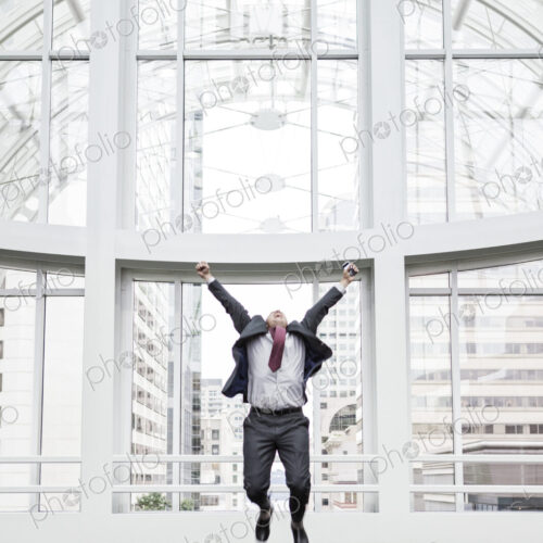A businessman jumping for joy in front of a large window in a convention center lobby.
