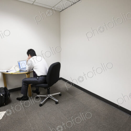 A businessman working alone at a small desk in an empty corner of an office.