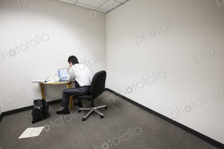 A businessman working alone at a small desk in an empty corner of an office.