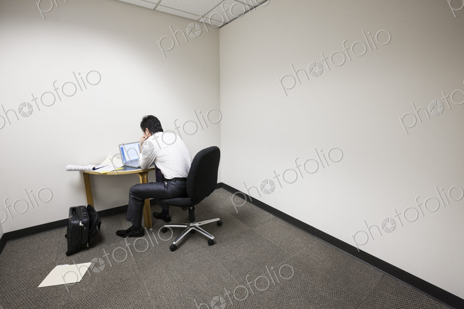 A businessman working alone at a small desk in an empty corner of an office.