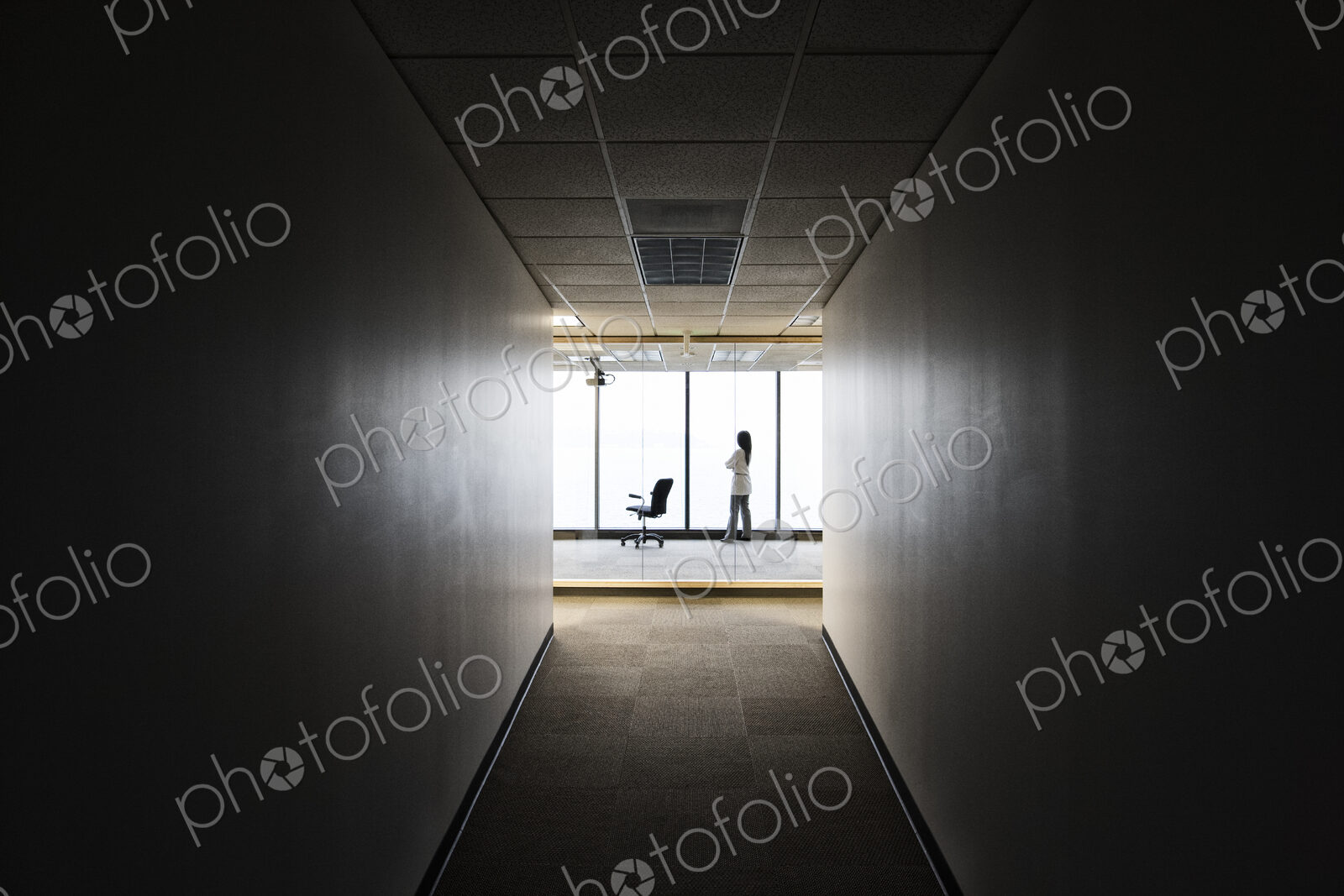 A businesswoman standing at a window at the end of a long hallway.