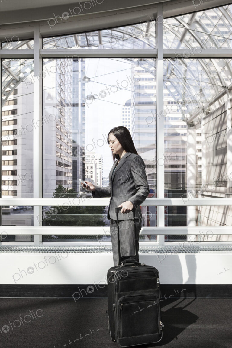 A businesswoman with a suitcase standing using a phone in a large open space under a…