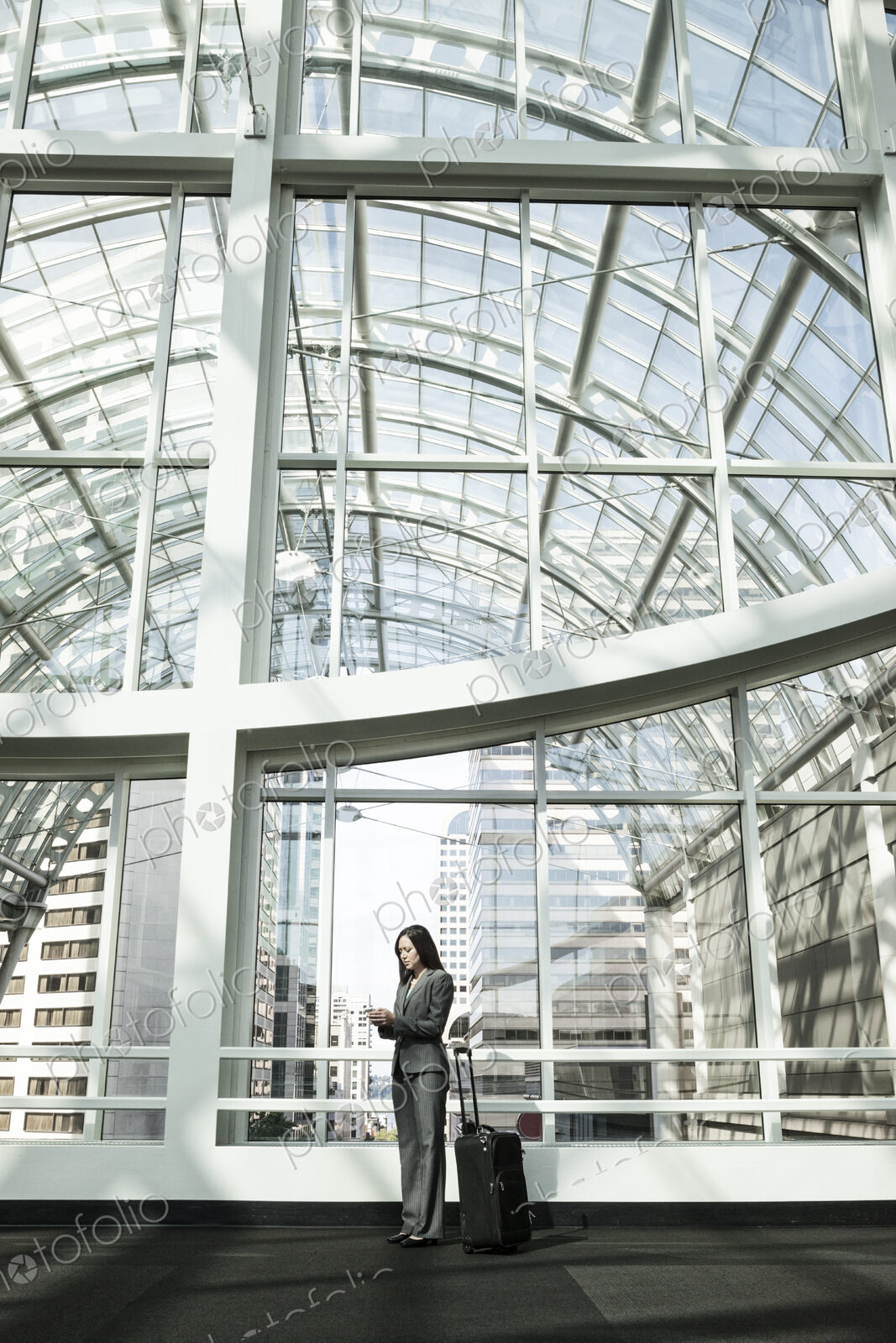 A businesswoman with a suitcase standing using a phone in a large open space under a…