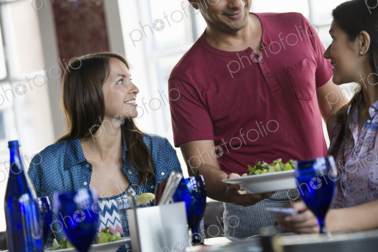 A cafe interior. A man in a waiter's apron serving a meal to two women at a table.