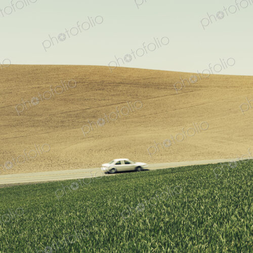 A car driving on am uphill slope, surrounded by farmland and lush, green fields of…