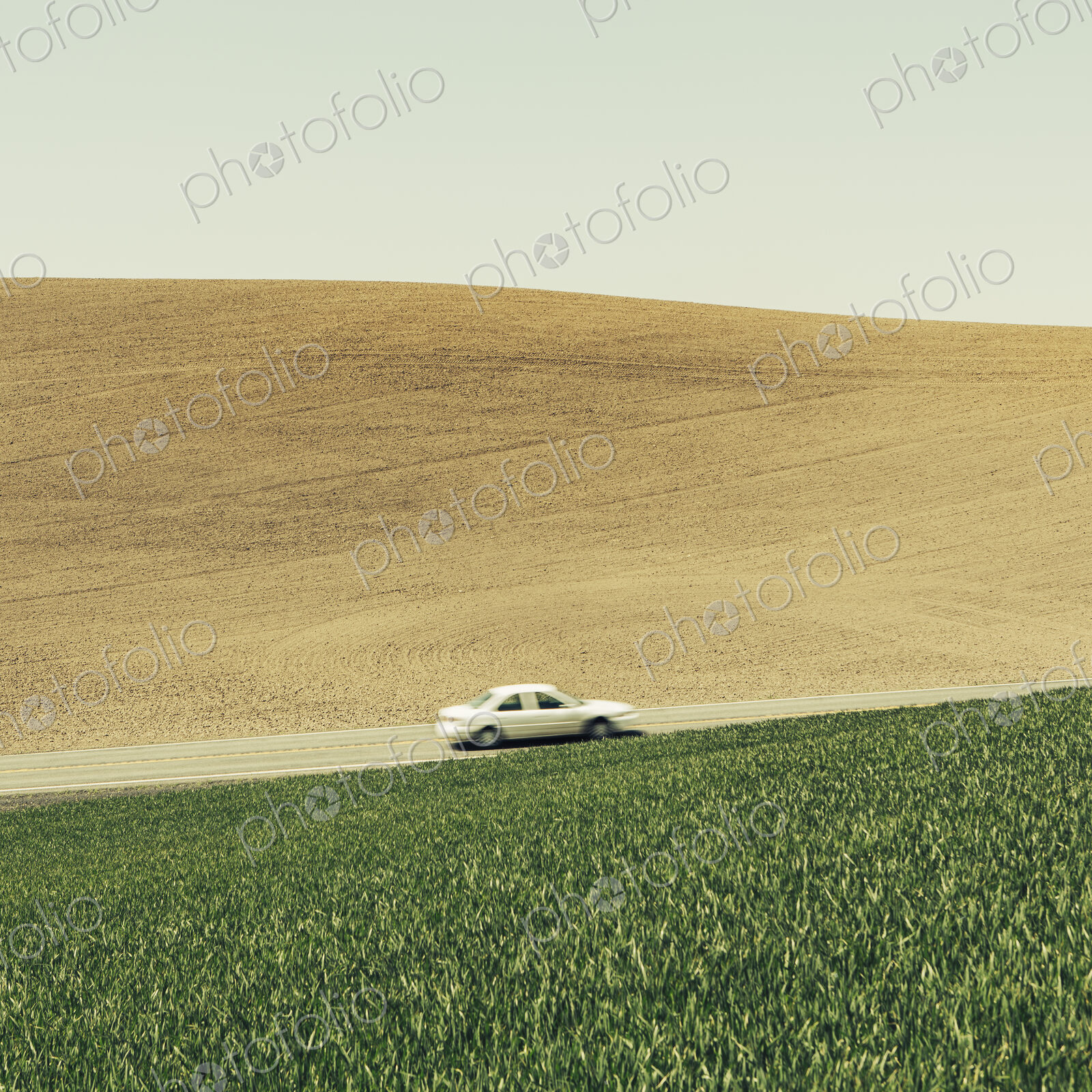 A car driving on am uphill slope, surrounded by farmland and lush, green fields of…