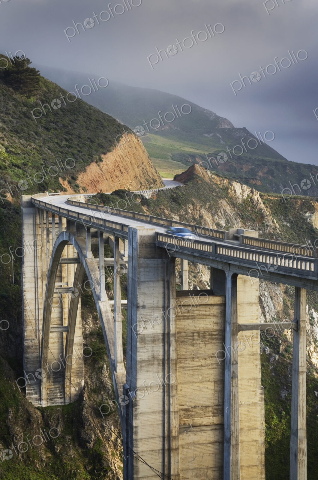 A car on a bridge on the Big Sur Pacific coastline, mountains and mist.