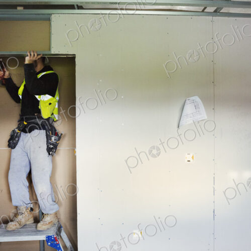 A carpenter in work clothes on a construction site, using an electric drill.