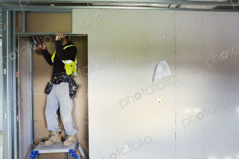 A carpenter in work clothes on a construction site, using an electric drill.