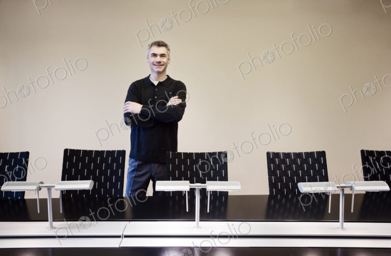 A casually dressed caucasian businessman standing behind an large office conference table.