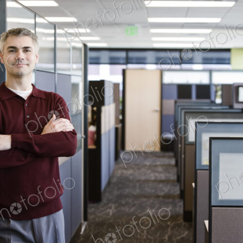 A casually dressed caucasian businessman standing next to his office in a room full of… A casually dressed caucasian businessman standing next to his office in a room full of…
