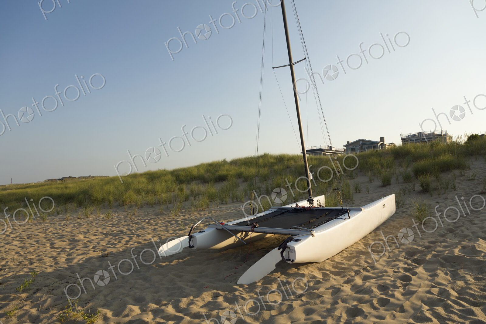 A catamaran sailing boat on the sand, on a beach, with houses in the distance.
