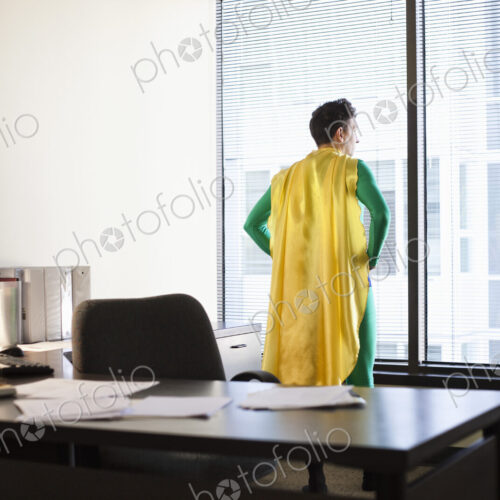 A caucasian businessman office super hero stands at his office window and ponders his…