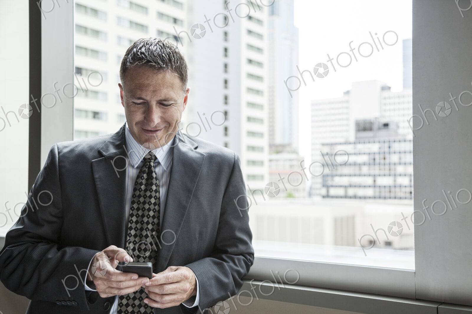 A caucasian businessman texting on his phone while standing next to a window.