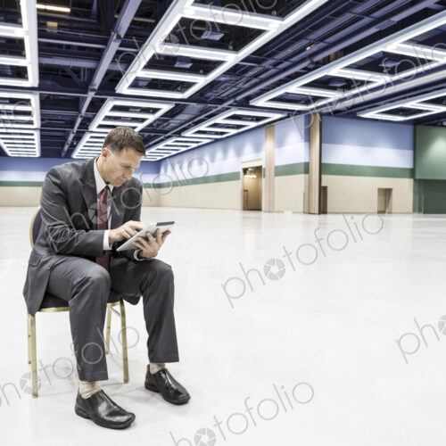 A caucasian businessman texting while sitting in a chair in the middle of a convention…