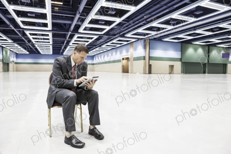 A caucasian businessman texting while sitting in a chair in the middle of a convention…