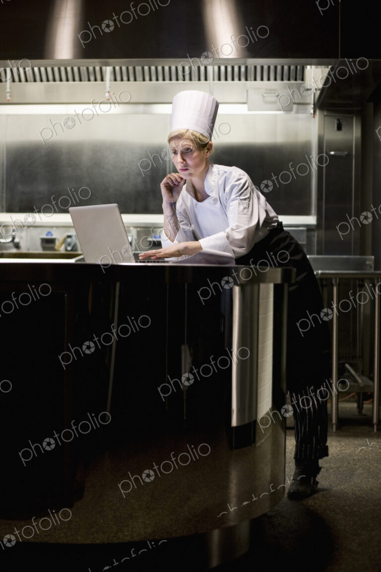 A caucasian female chef working on a laptop computer after hours in a commercial kitchen.