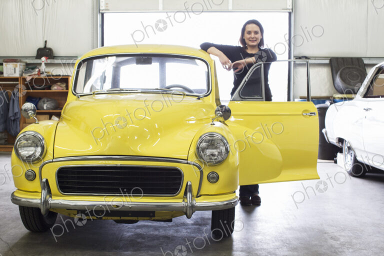 A caucasian female stands next to her old car in a classic car repair shop.