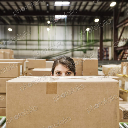 A caucasian female warehouse worker behind a cardboard box in a distribution warehouse.