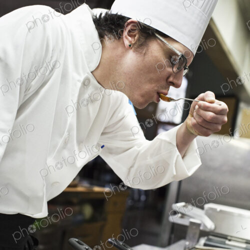 A caucasian male chef tastes a sample of the sauce he is making in a commercial kitchen,