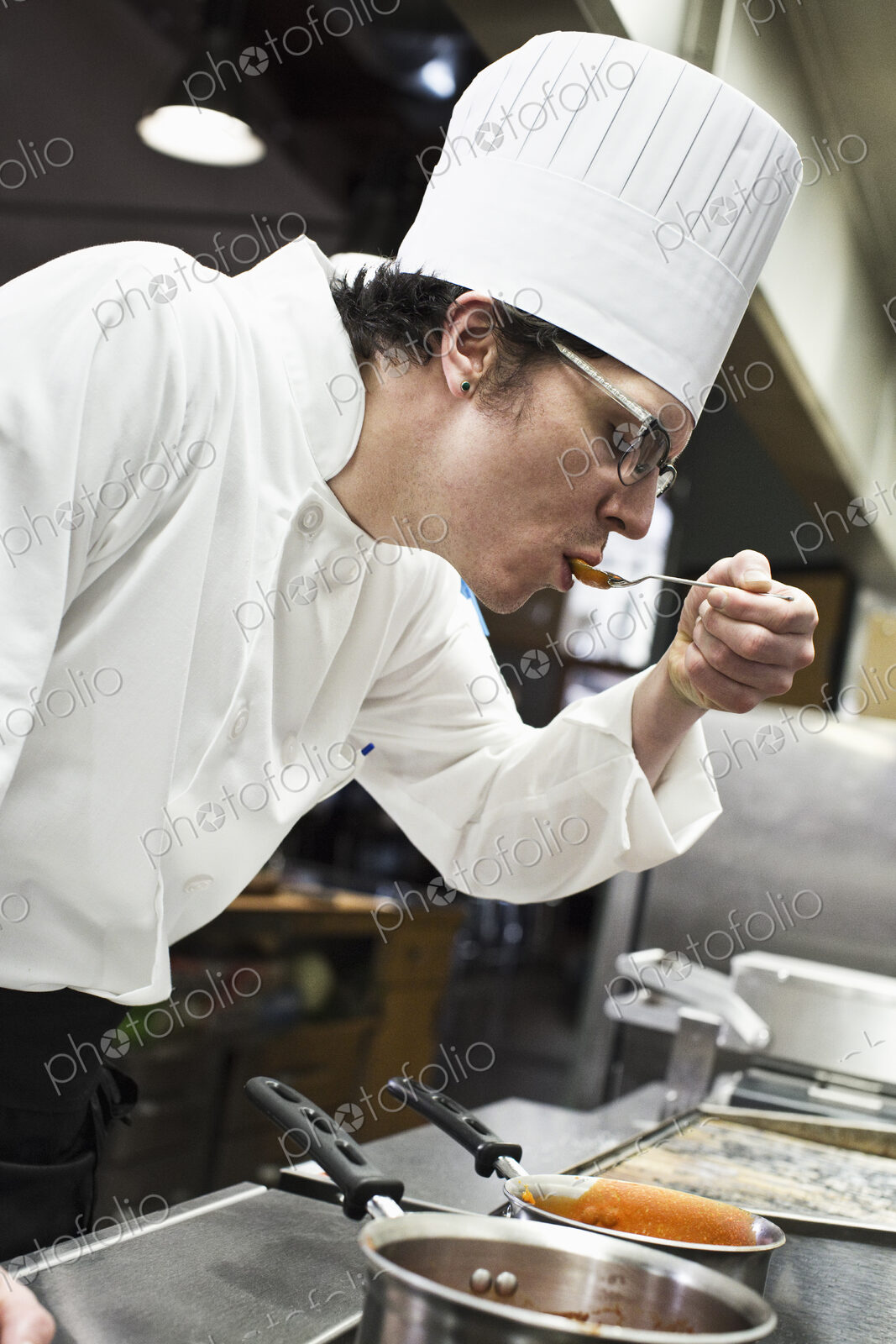 A caucasian male chef tastes a sample of the sauce he is making in a commercial kitchen,