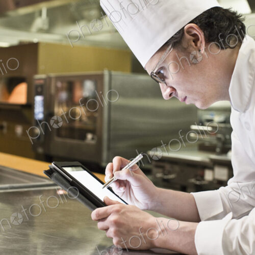 A caucasian male chef working on a notebook computer in a commercial kitchen.