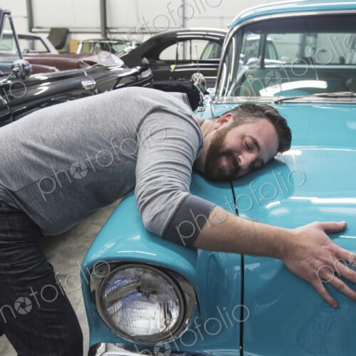 A caucasian male hugging the hood of his old sedan in a classic car repair shop.