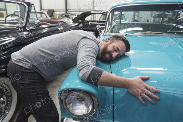 A caucasian male hugging the hood of his old sedan in a classic car repair shop.