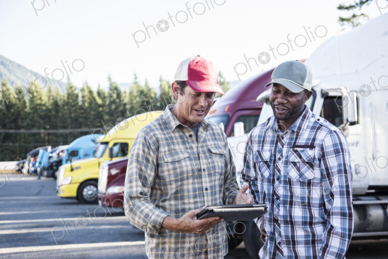 A caucasian man and a black man truck driving team together in a truck stop parking lot.
