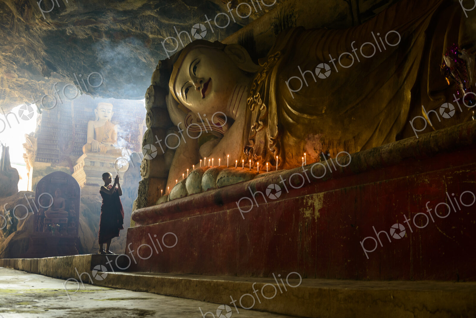 A cave temple, a large reclining Buddha statue, lit candles and a monk praying.