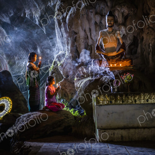 A cave temple with Buddha statues, lit candles and two girls kneeling and praying. A cave temple with Buddha statues, lit candles and two girls kneeling and praying.