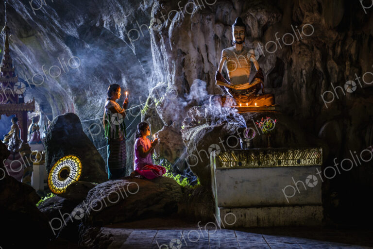 A cave temple with Buddha statues, lit candles and two girls kneeling and praying.