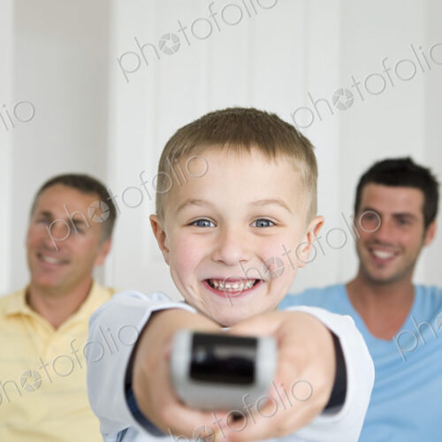 A child, a boy holding a television remote control. Two men on the sofa behind him. A child, a boy holding a television remote control. Two men on the sofa behind him.