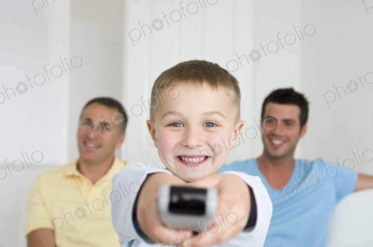 A child, a boy holding a television remote control. Two men on the sofa behind him.
