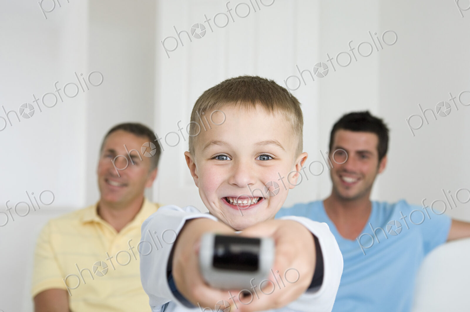 A child, a boy holding a television remote control. Two men on the sofa behind him.