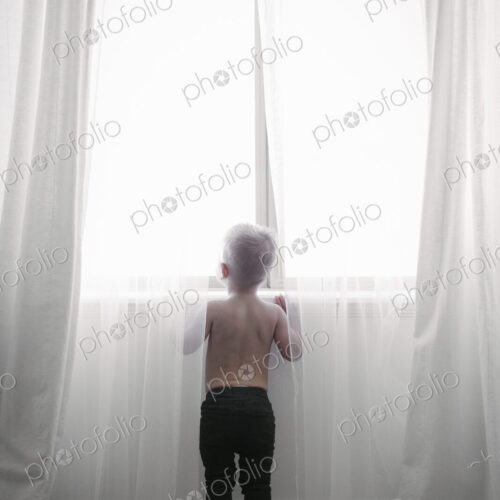 A child standing at a window looking out through the net curtains. Back view. A child standing at a window looking out through the net curtains. Back view.