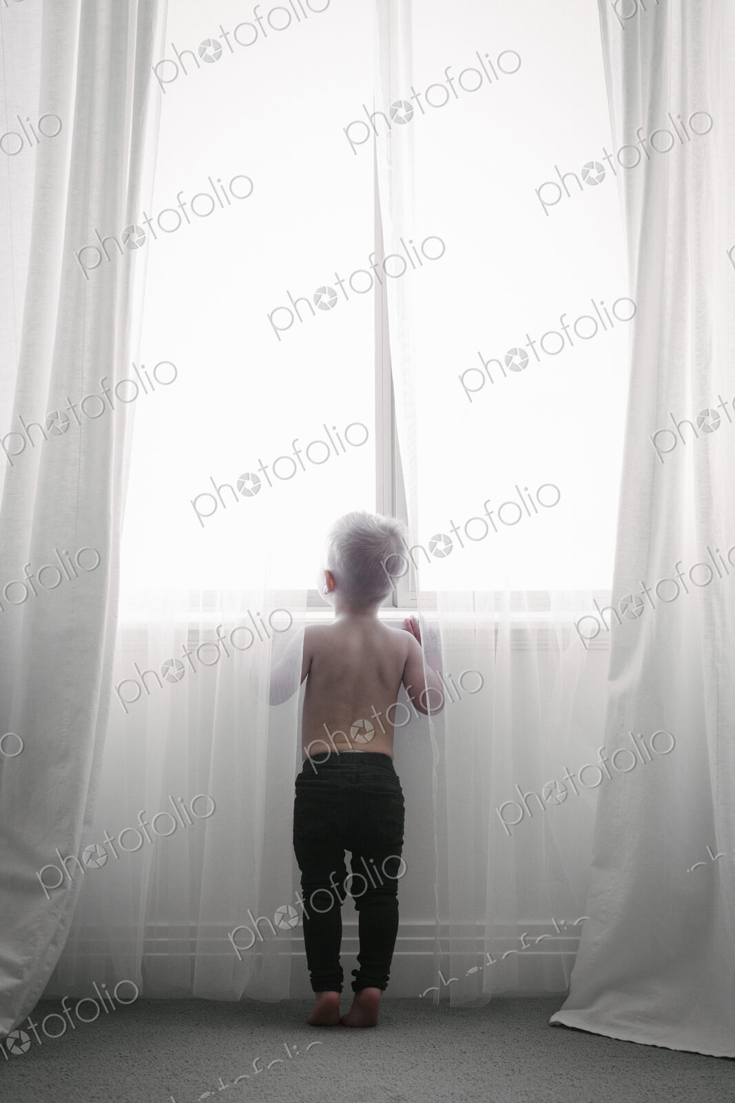 A child standing at a window looking out through the net curtains. Back view.