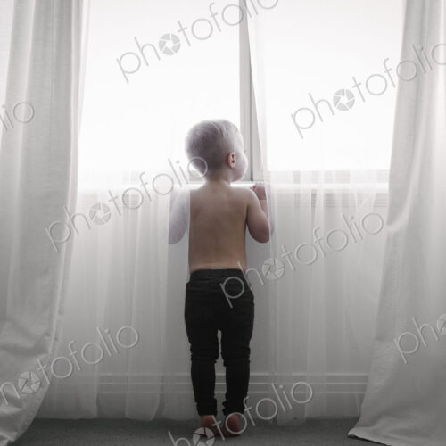 A child standing at a window looking out through the net curtains. Back view.