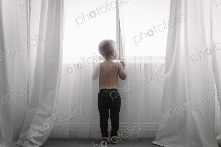 A child standing at a window looking out through the net curtains. Back view.