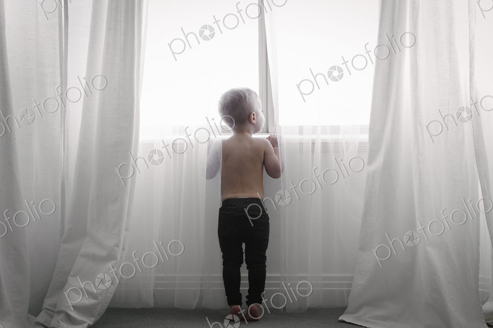 A child standing at a window looking out through the net curtains. Back view.