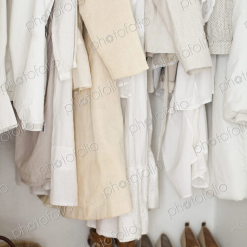 A closet with cream and white clothes, jackets, shirts and tunics hanging up. A row of…