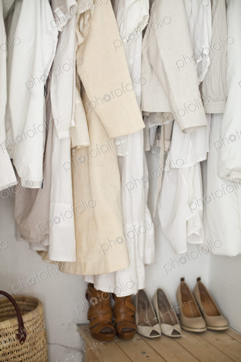 A closet with cream and white clothes, jackets, shirts and tunics hanging up. A row of…